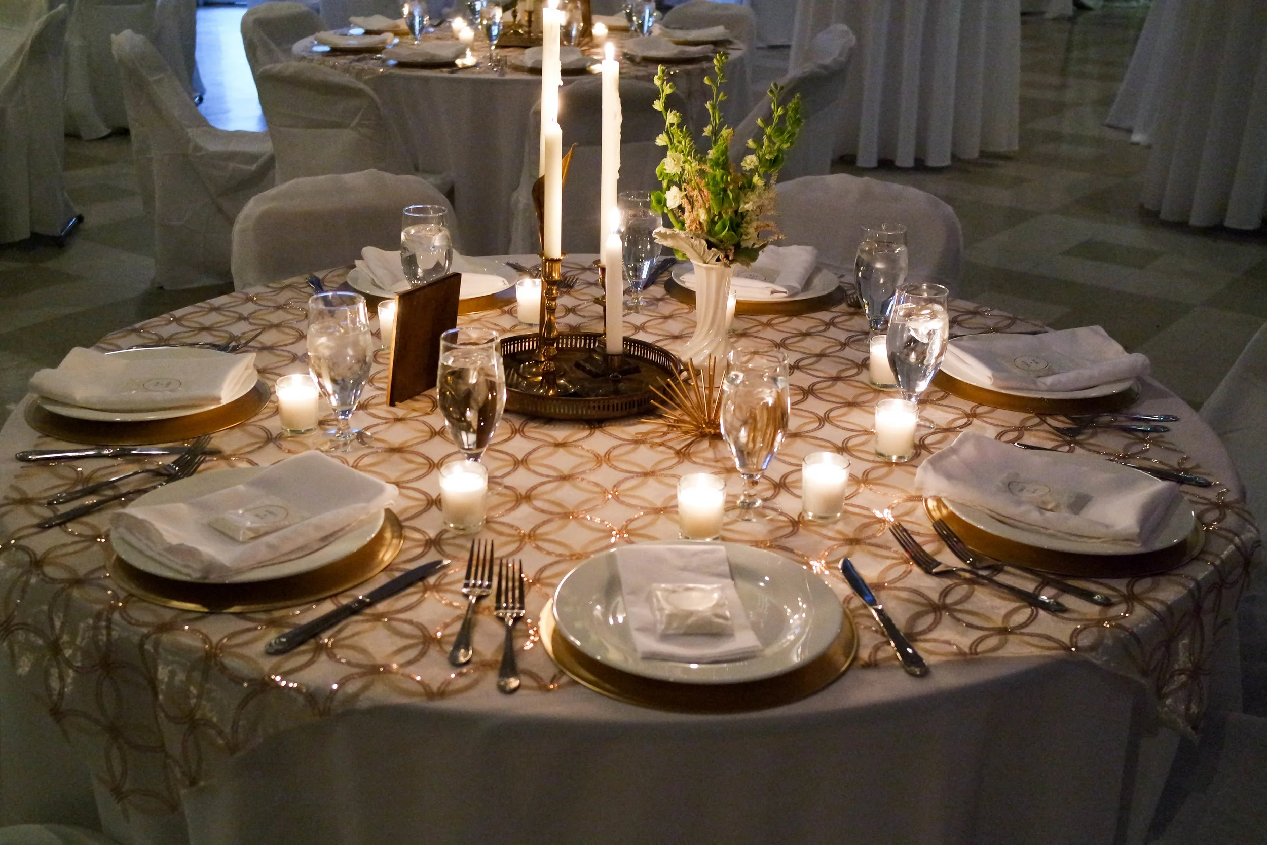 A wide interior view of an elegant banquet hall set for a formal evening, round tables dressed in white linens, polished gold-rimmed plates, tall white taper candles, and low floral centerpieces, warm amber and cream tones, hushed atmosphere.